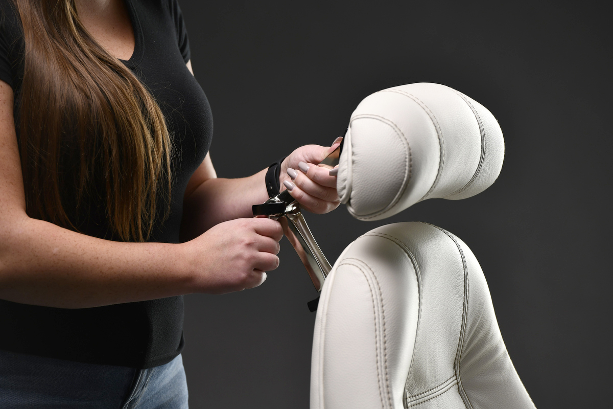Lifeform.Chairs.D2.00158 A woman with long brown hair adjusts the chrome mechanism of a padded headrest on an off-white, leather-style ergonomic chair against a dark grey background.