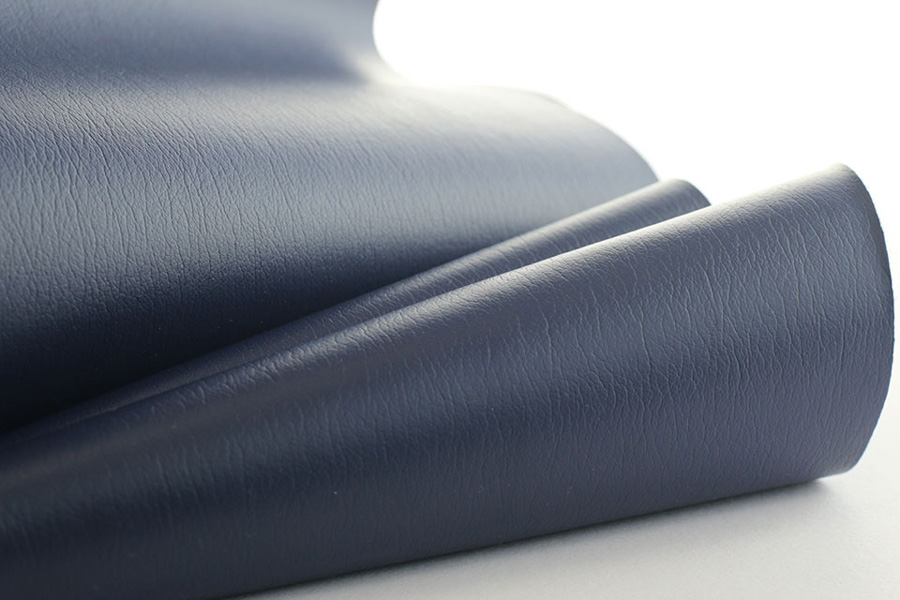 Close-up of folded navy blue leatherette with a fine, natural grain and smooth surface, creating soft curves and shadows that highlight the material’s texture. A small red square logo reading “Uf Ultrafabrics” appears in the bottom-right corner, indicating the brand.