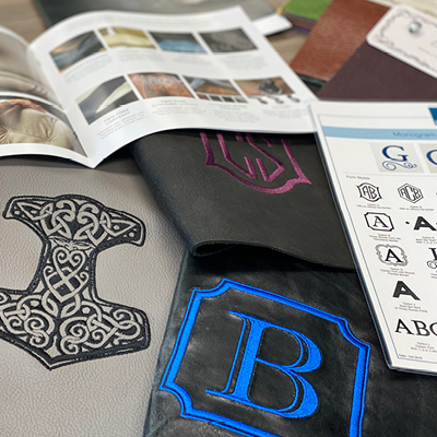 Assorted leather samples featuring embroidered monogram designs, displayed alongside catalog pages showing fonts and customization options.