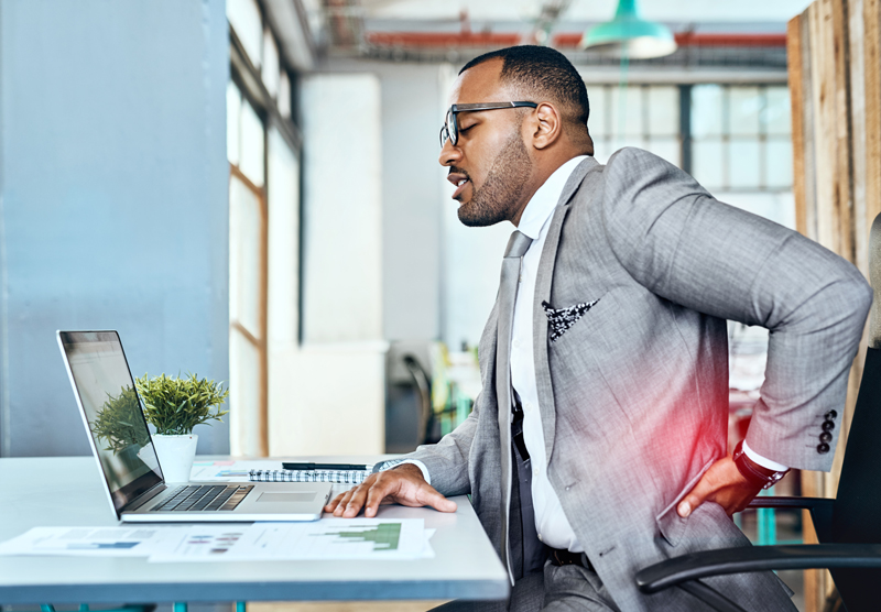 A man in a grey suit sits at a desk and holds his lower back in apparent discomfort while working on a laptop.