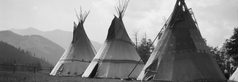 Three traditional Stoney First Nations tipis standing in a grassy field with mountains in the background.