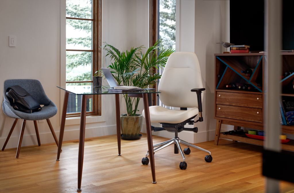 White ergonomic office chair with adjustable armrests on casters next to glass-top table with potted plants in modern home office with wooden flooring and large windows.