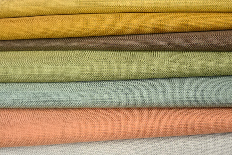 Stack of folded woven fabric swatches arranged horizontally, showing textured surfaces in warm and muted colours including mustard yellow, brown, olive green, pale blue, soft peach and light grey.