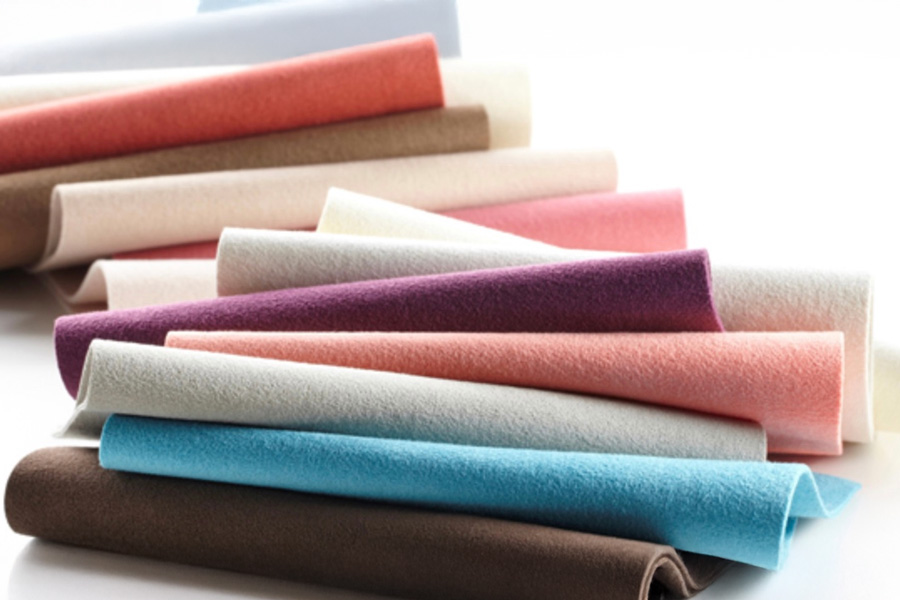 Several soft, felt-like fabric swatches rolled and layered in a loose fan across a white surface, showing a range of colours—brown, turquoise blue, pale grey, peach, pink, plum and cream—with a slightly fuzzy texture and clean, straight edges, presented as material and color samples.