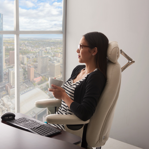 Woman with glasses sitting in a beige ergonomic office chair at a high-rise desk, holding a cup whilst looking out of a floor-to-ceiling window overlooking a cityscape. The chair features a headrest and lumbar support, demonstrating ergonomic design for extended work periods.